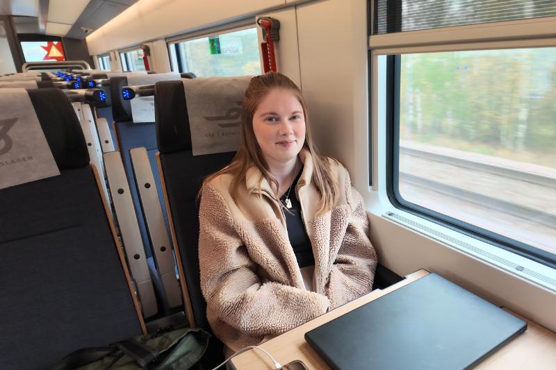 Kvinna sitter ombord på tåget, framför henne på bordet finns en dator. Beige jacka och brunt hår.