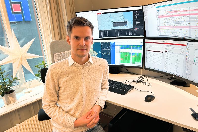Man står vid ett skrivbord, i bakgrunden finns massa datorskärmar. Emil arbetar som trafikledare. Brunt hår, vit tröja.